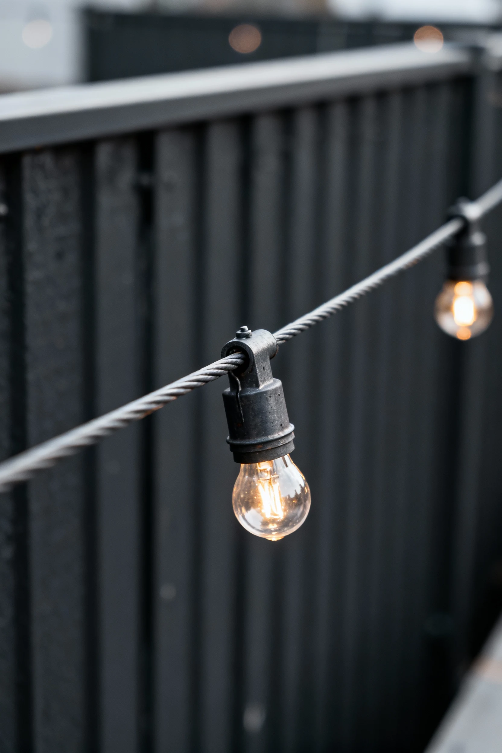 closeup steel guide wire holding string lights, charcoal fence